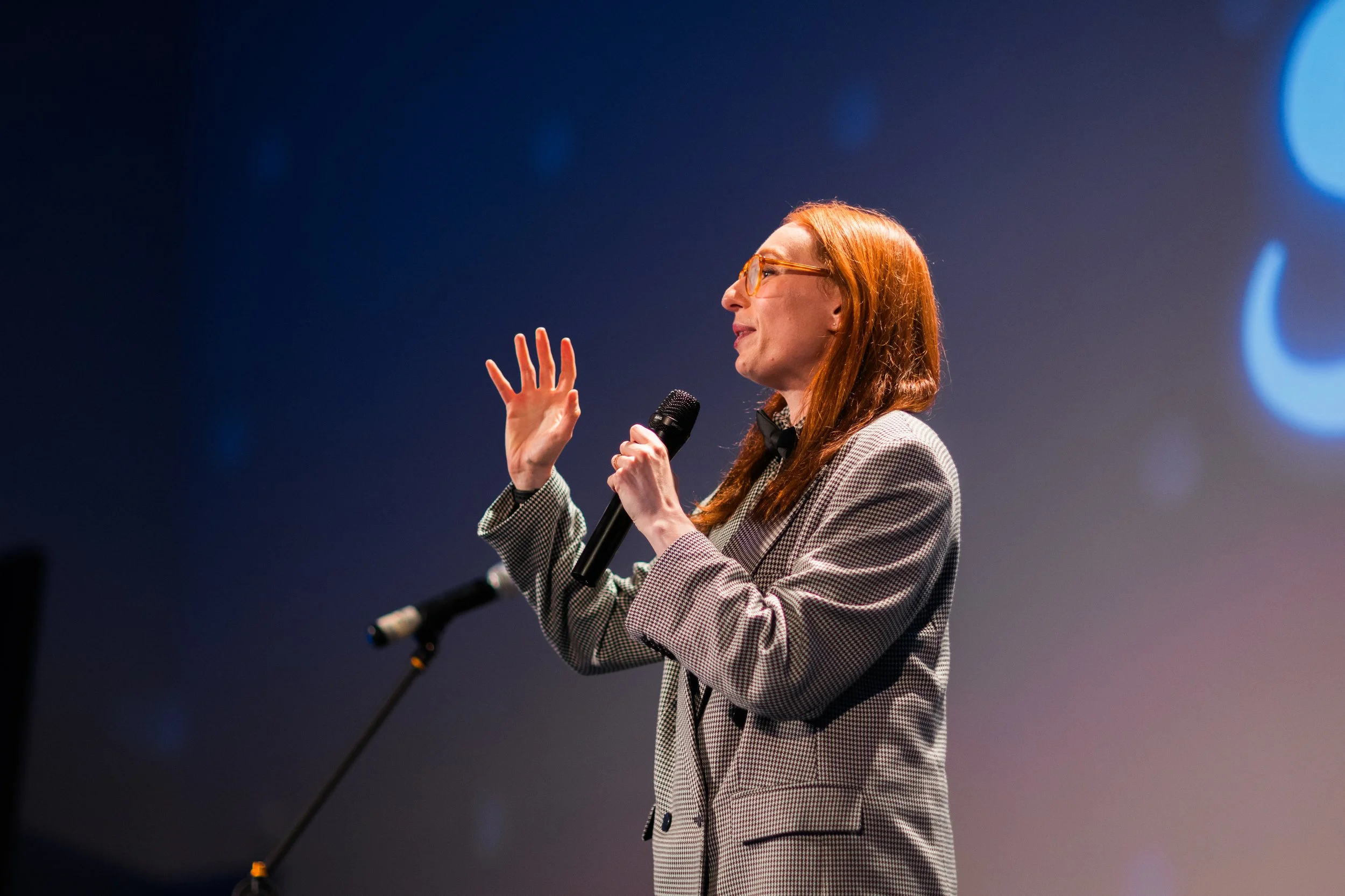 A woman with red hair and glasses, wearing a houndstooth blazer, speaking into a microphone on stage with a blue background.