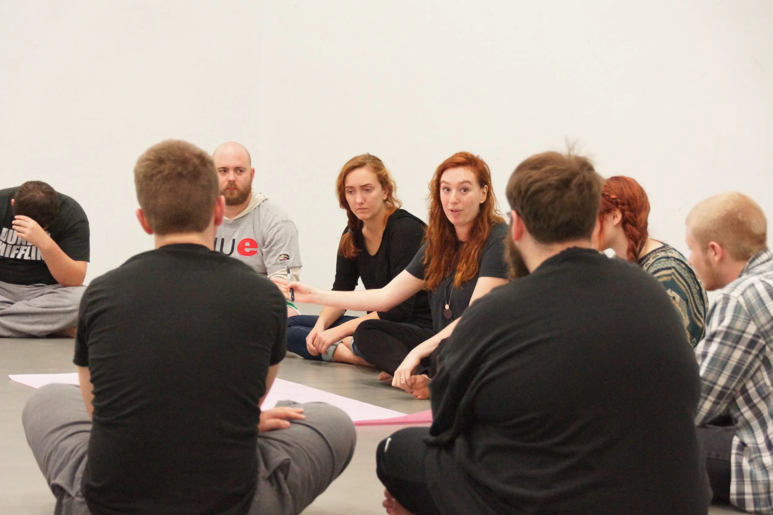 Group of young adults sitting on the floor in a circle, participating in a discussion in a room with a plain white wall.