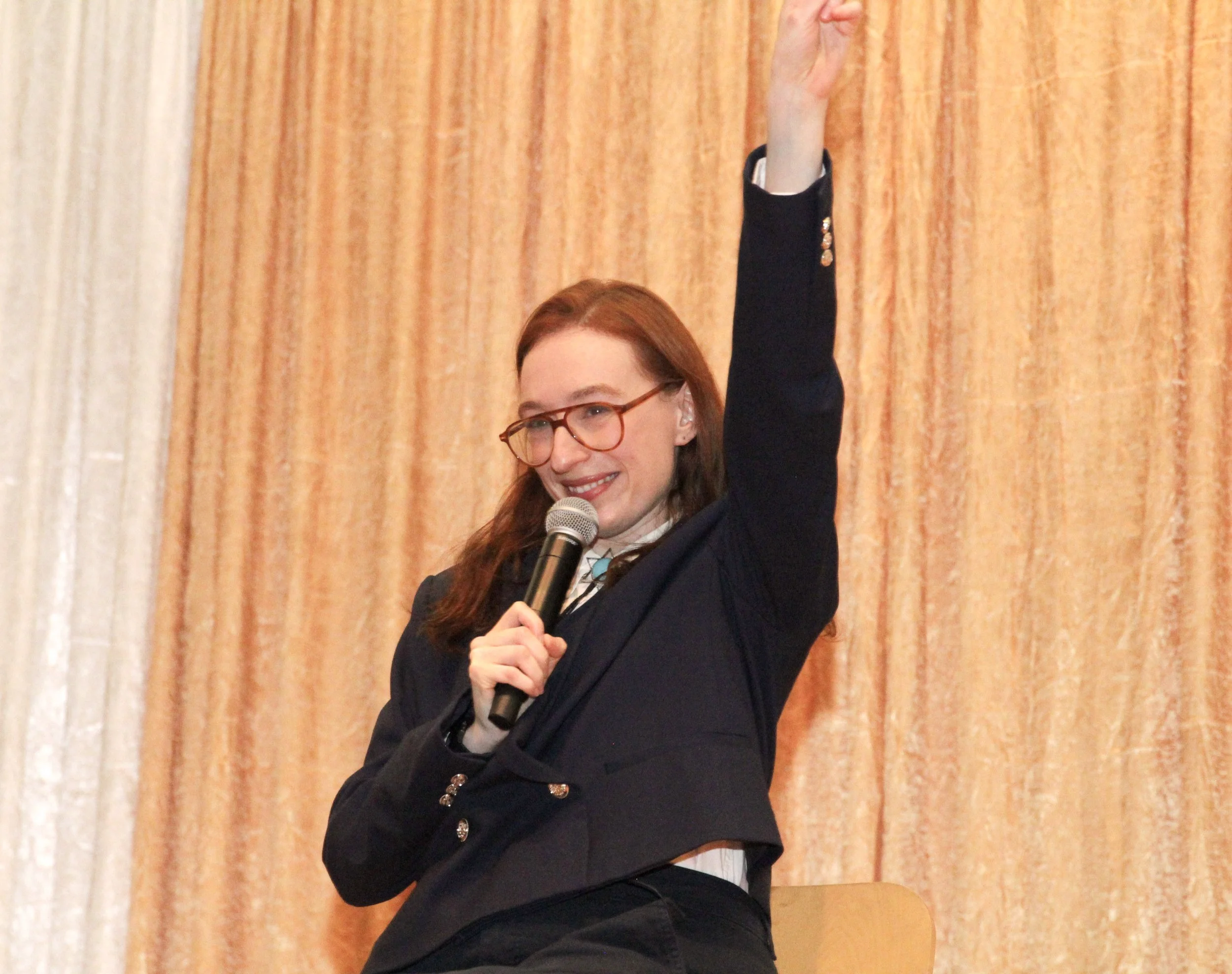 A woman with red hair and glasses is smiling, holding a microphone, and raising her right arm with her hand clenched in a fist. She is dressed in a dark blazer with decorative buttons and a white shirt, standing in front of a beige curtain.