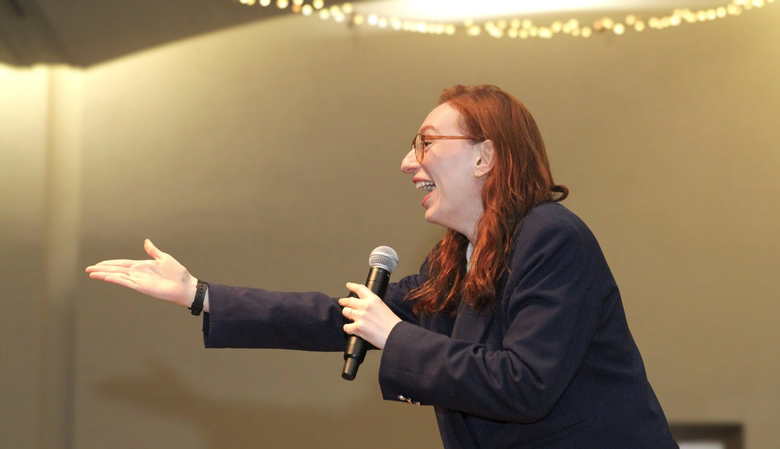 A woman with red hair and glasses holding a microphone, gesturing with her left hand while speaking and smiling during a presentation or speech.