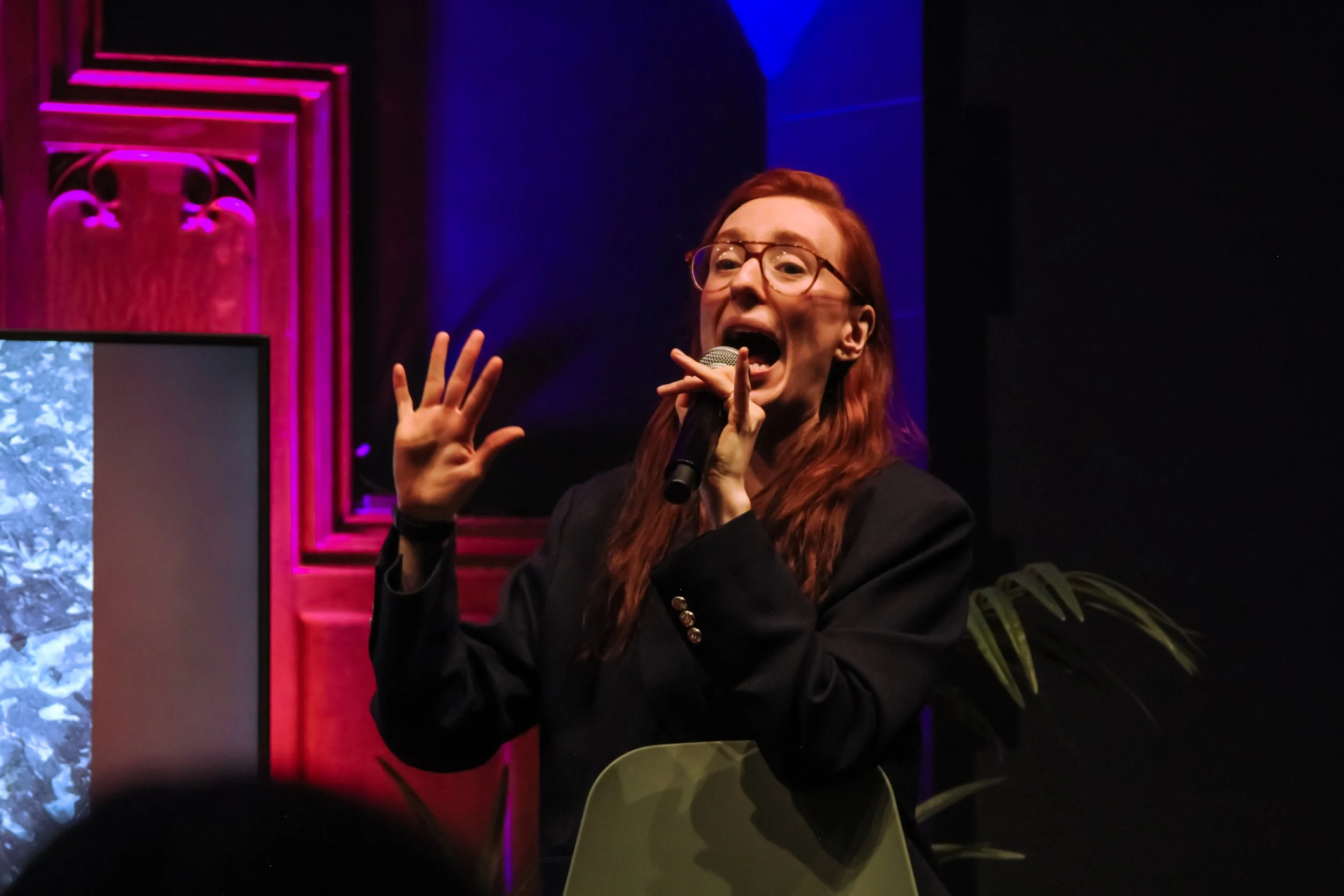 A woman with red hair and glasses holding a microphone and speaking or singing, with colorful lighting in the background, including pink and blue lights.