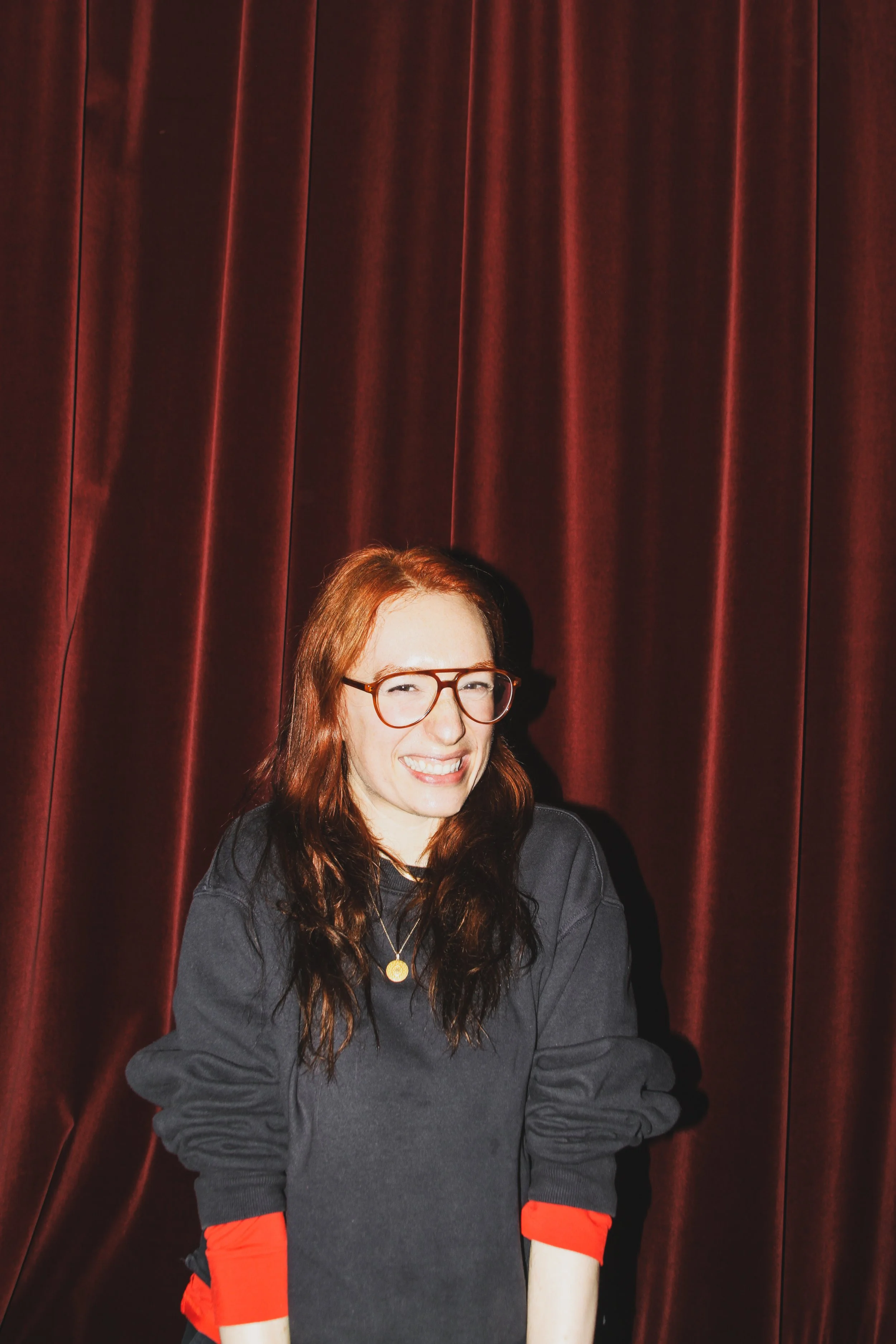 A woman with long red hair, glasses, and a black sweatshirt standing in front of a red curtain, smiling.