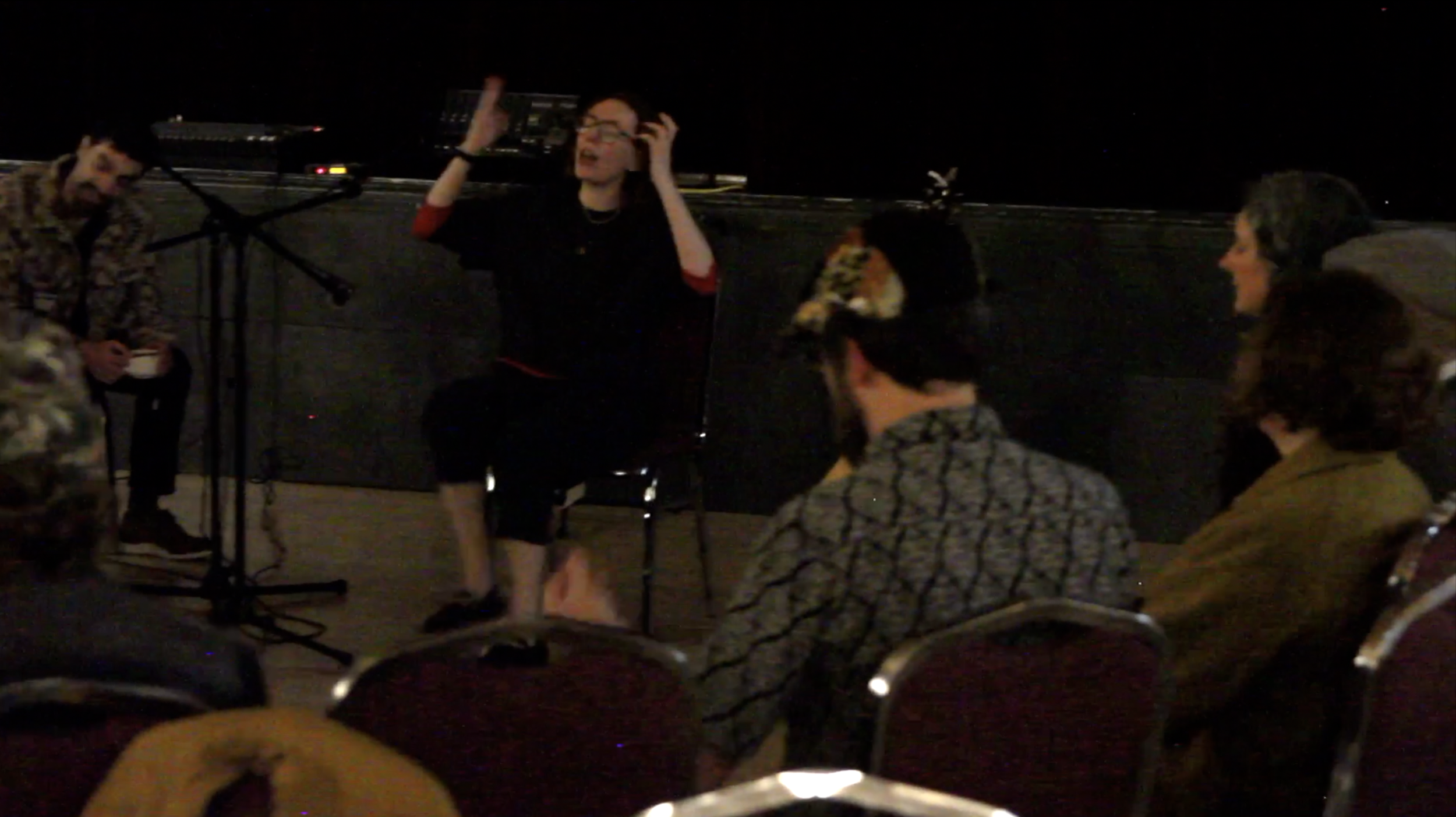 Group of people gathered on a rooftop at night, engaging in a discussion or presentation with a person speaking and gesturing, surrounded by chairs and microphones.