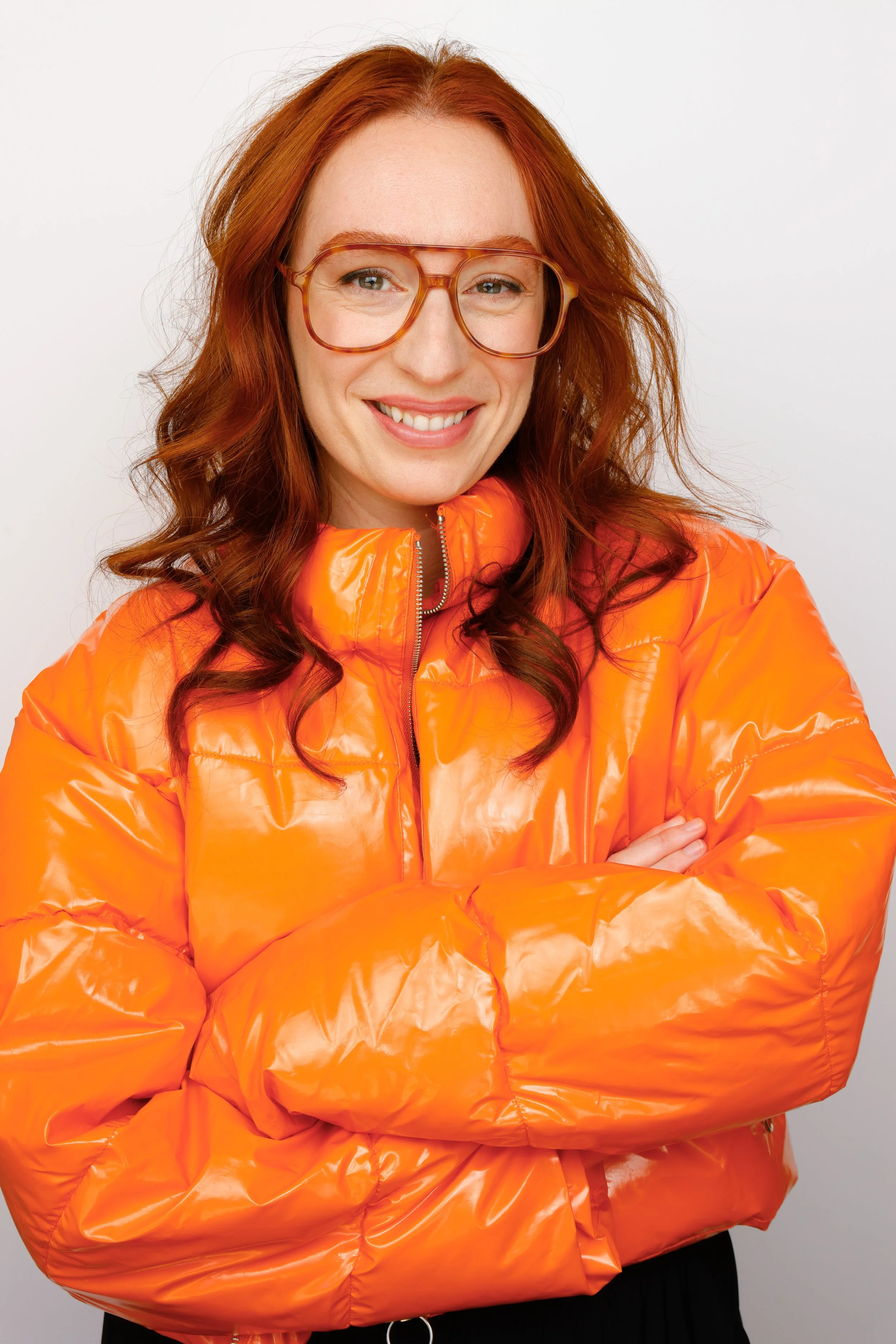 A woman with red hair wearing large glasses and a shiny orange puffer jacket smiling with arms crossed.