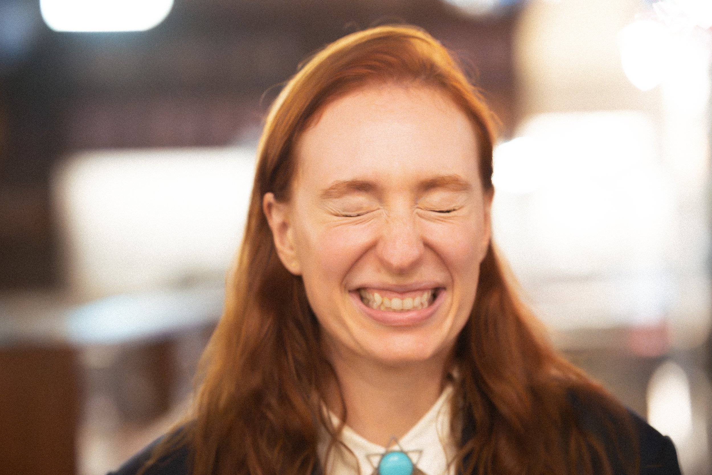 A woman with red hair and a bright smile, eyes closed, in an indoor setting with blurred background of lights.