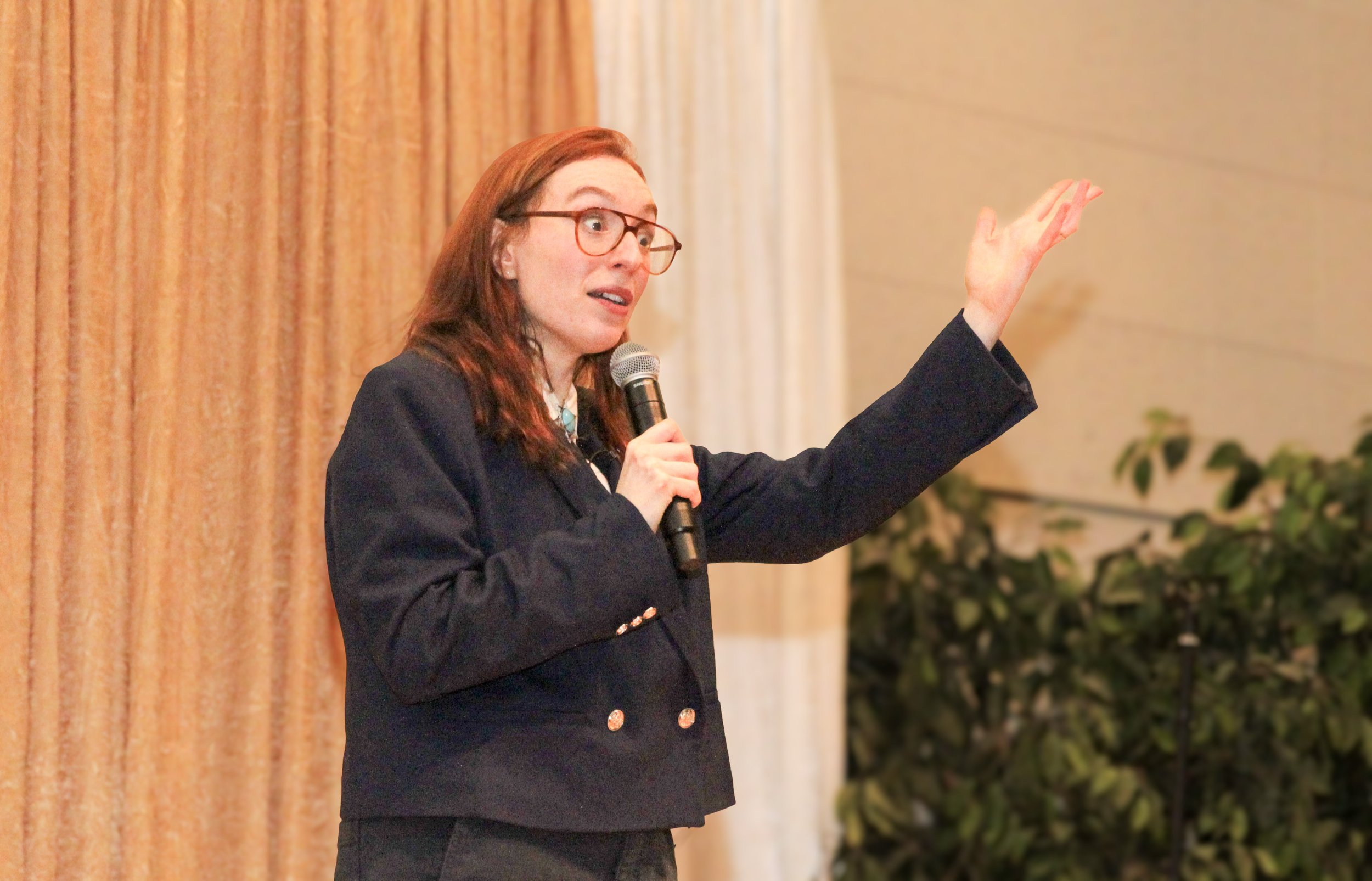A woman with red hair and glasses giving a presentation, holding a microphone, and gesturing with her left hand in a meeting or conference room.