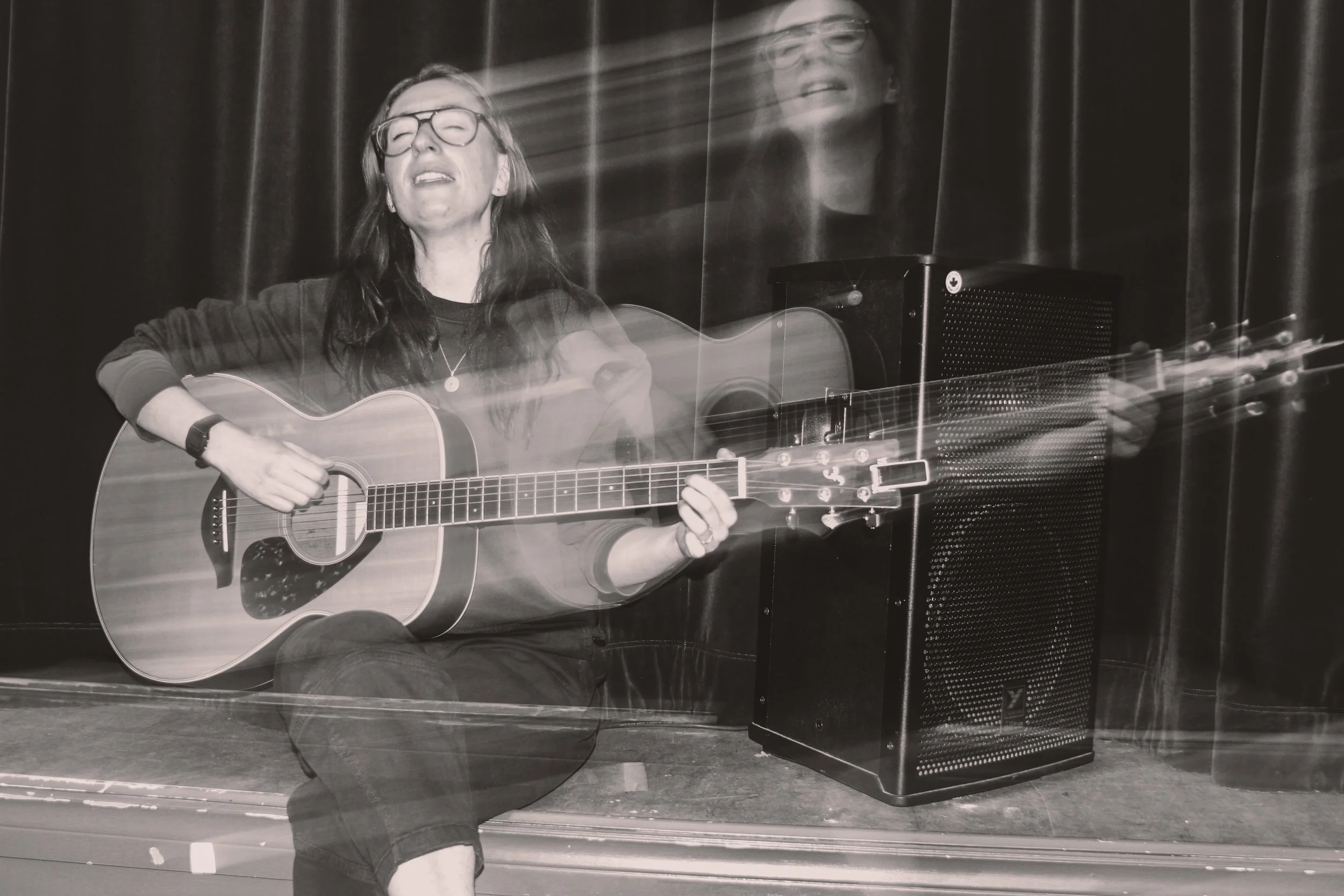 Black and white photo of a woman playing an acoustic guitar on stage, with a microphone in front of her and a speaker on her right.