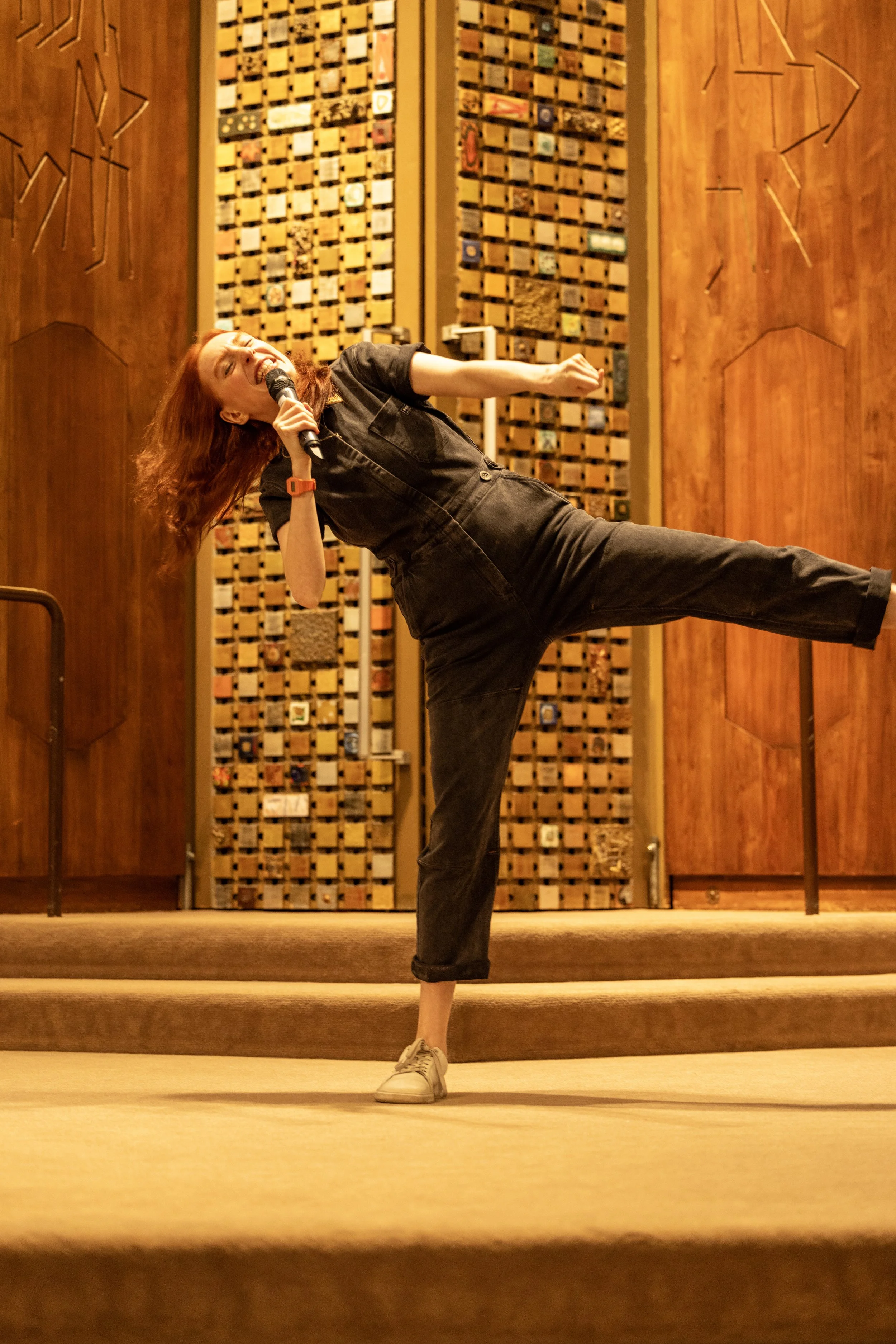 A woman singing on stage, holding a microphone and performing a dance move, in front of a wooden background with a decorative pattern.
