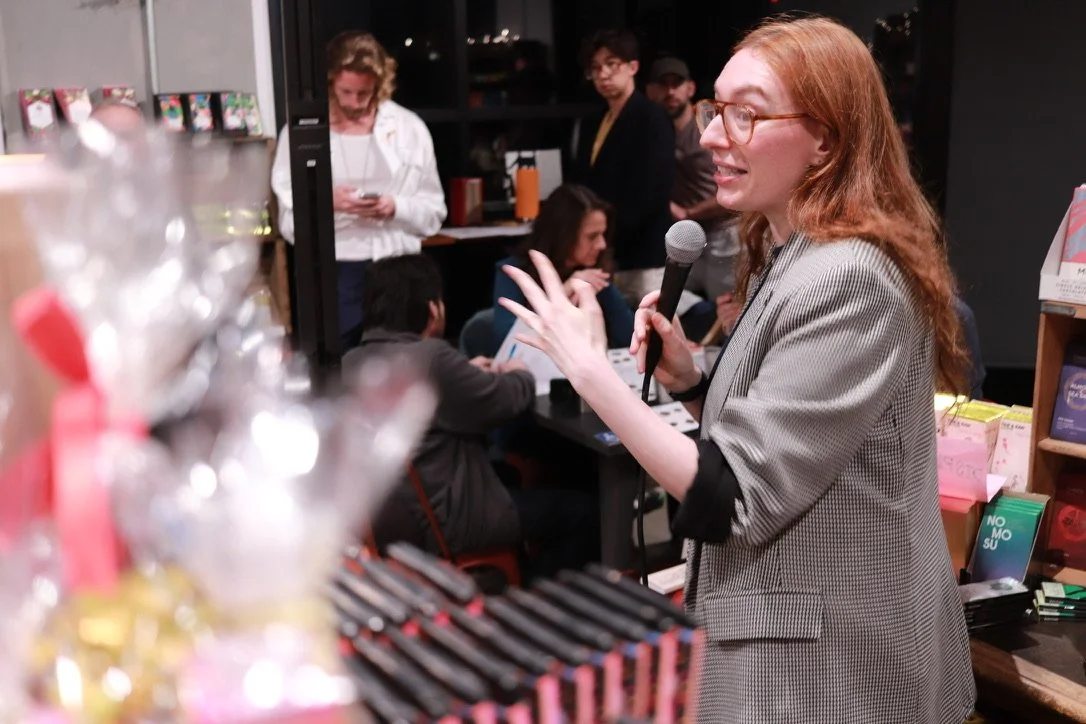 A woman with red hair and glasses holding a microphone speaking to a group of people in a bookstore or cafe, with books and snacks in the foreground.