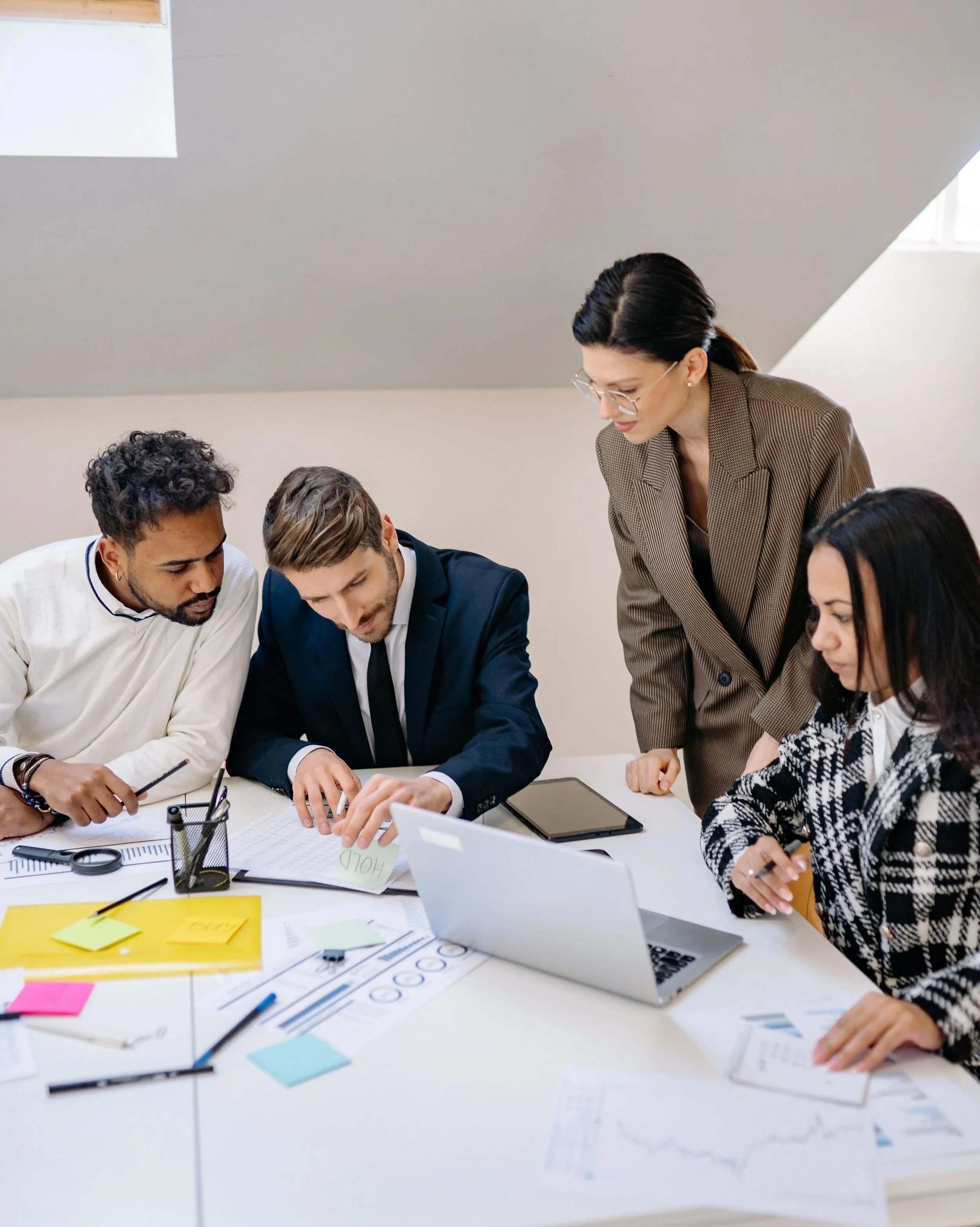 A diverse group of people working together at a table in an office. They are discussing documents and using electronic devices, with office supplies and papers scattered on the table.