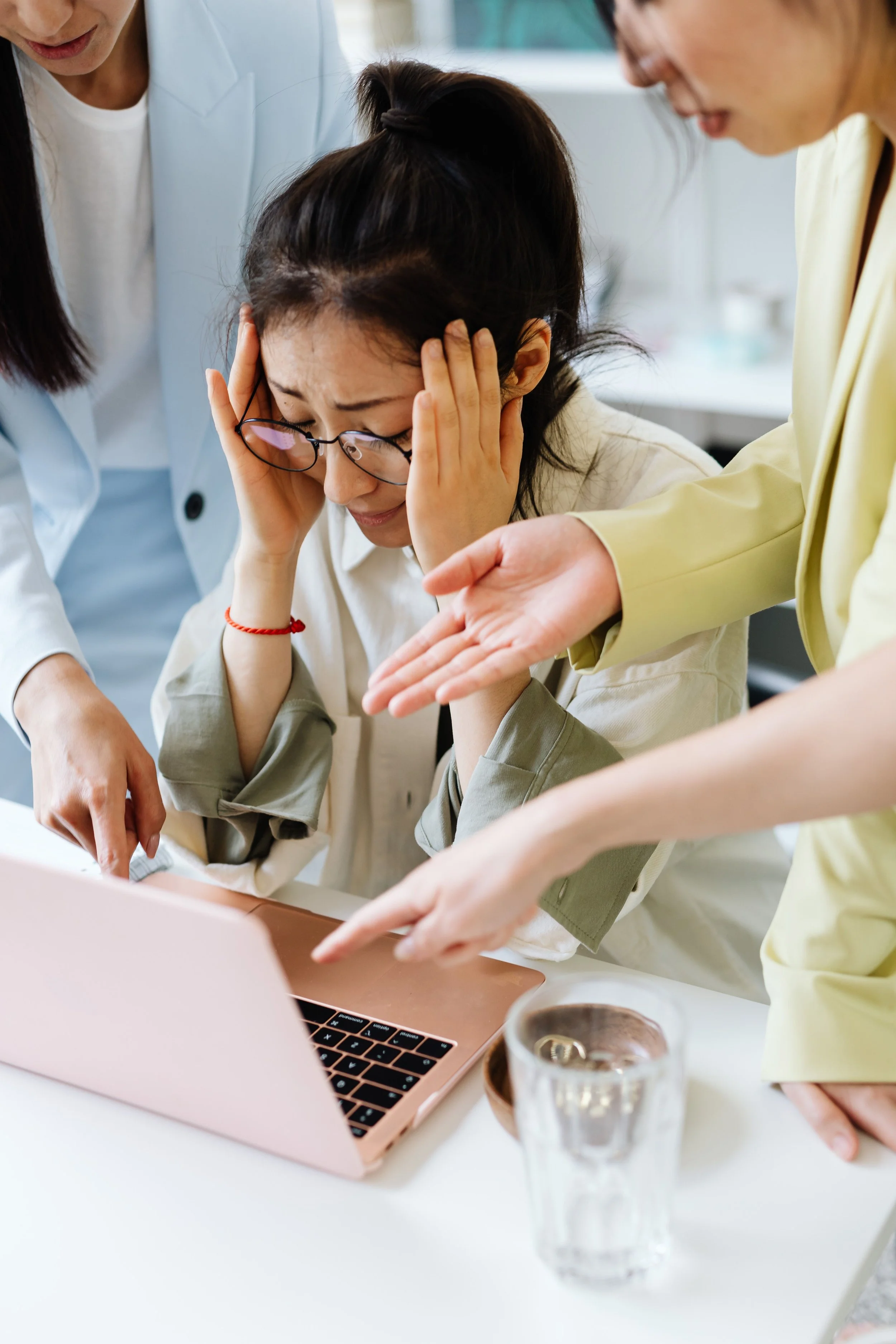 A woman with dark hair and glasses looks distressed while holding her head during a discussion with three other women around her, in a well-lit room with a laptop and glasses of water on the table.