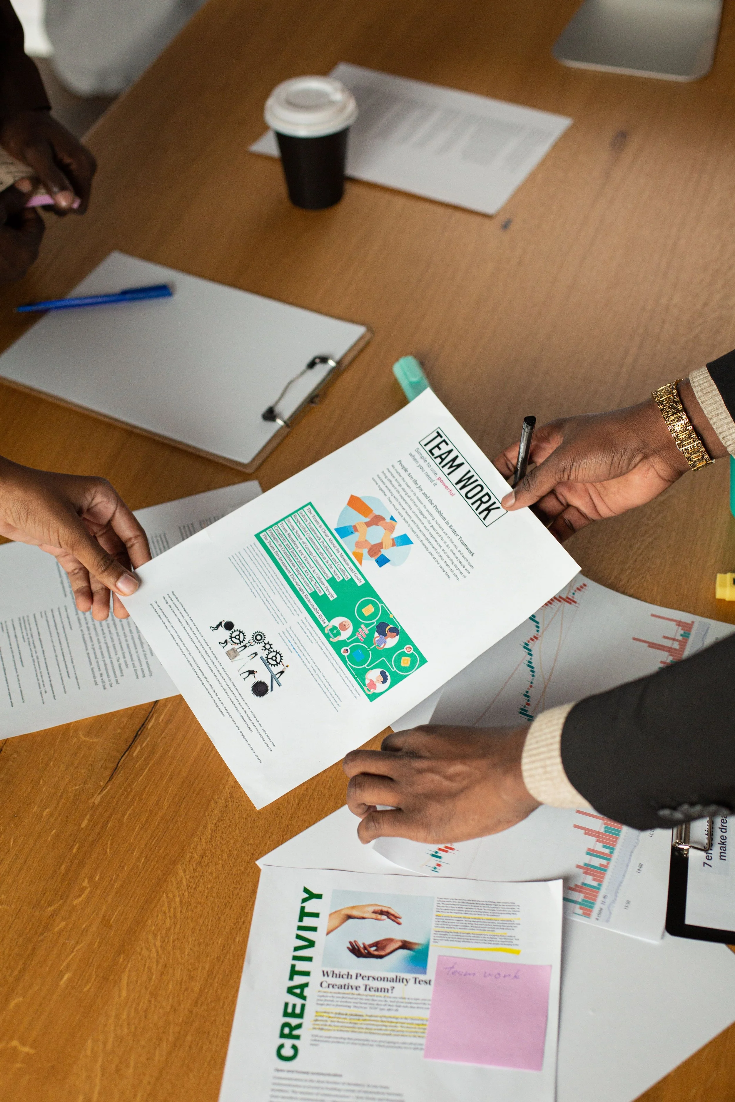 People working on a table with various printed documents, charts, and a clipboard, discussing teamwork and productivity.