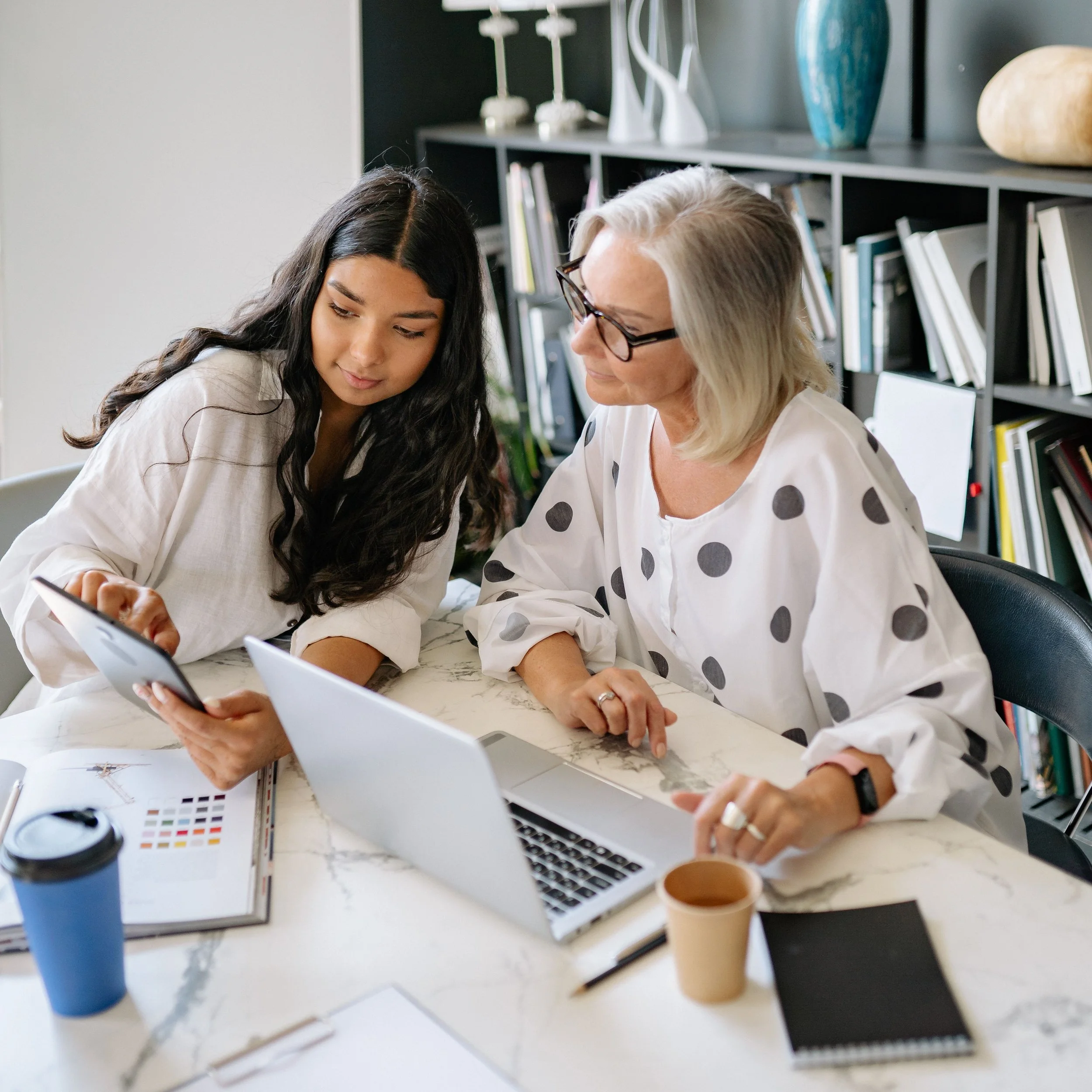 Two women sitting at a table looking at a tablet in a modern office space with bookshelves, vases, and coffee cups on the table.
