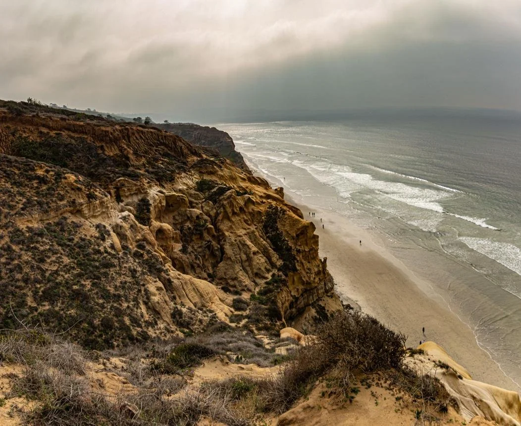 A view of a rugged coastline with cliffs and a sandy beach, with ocean waves and cloudy sky in Two women talking on a beach in Carlsbad, California.