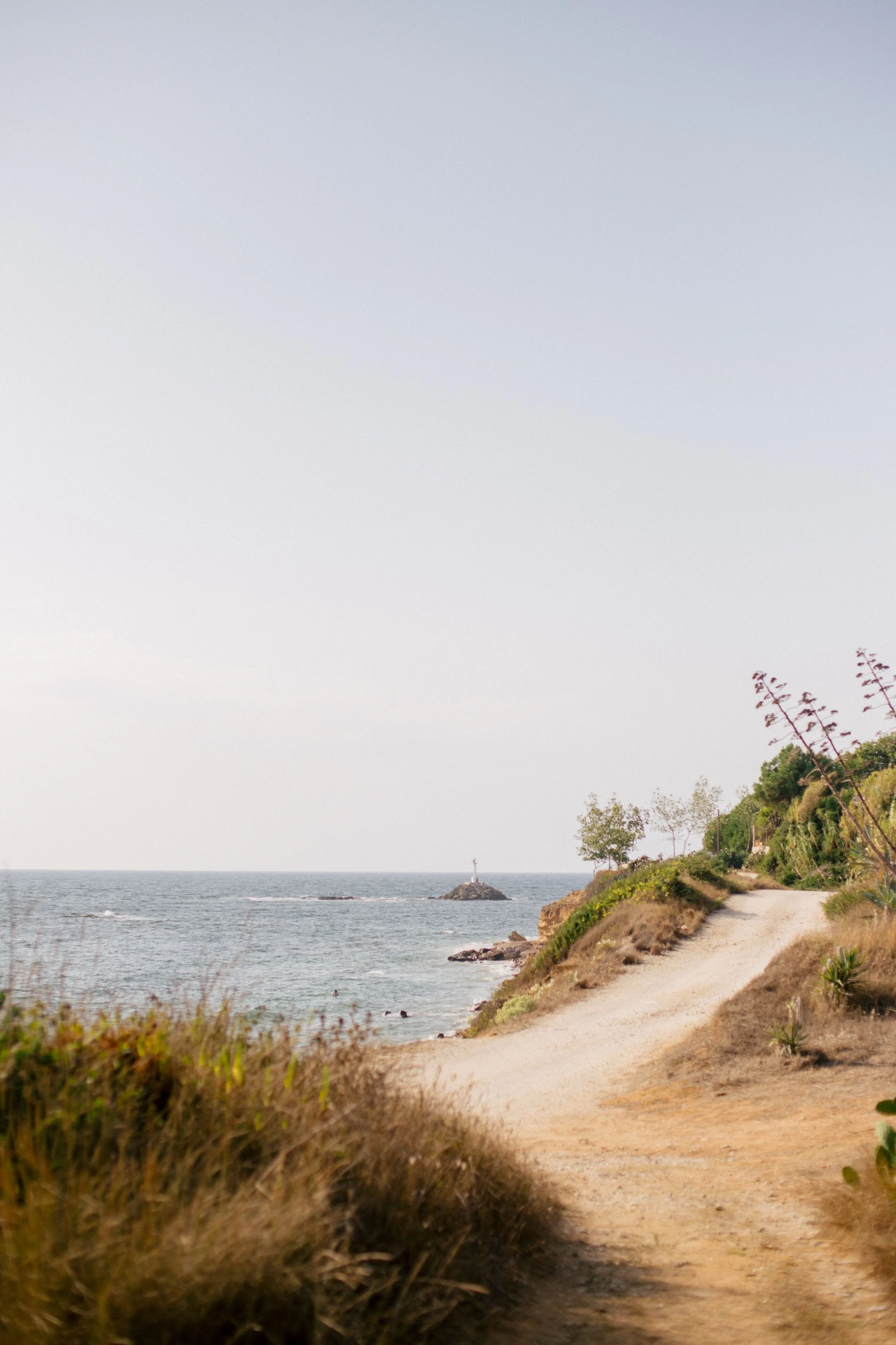 A coastal scene with a dirt pathway along a hillside, leading towards the ocean. The coastline features sparse vegetation and small trees, with a small lighthouse on a rock outcrop in the water.