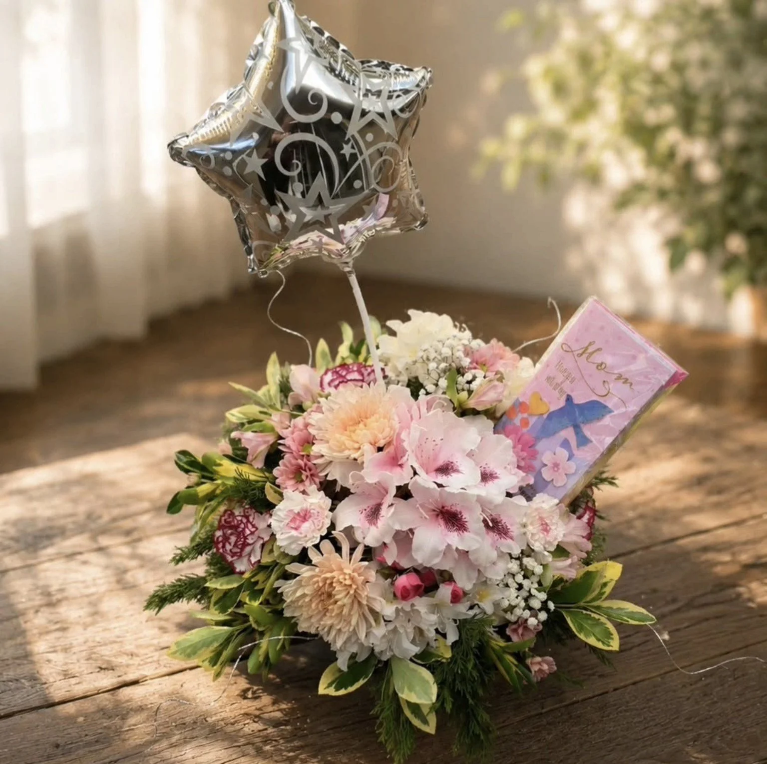 A bouquet of pink and white flowers with green leaves, a silver star-shaped balloon with stars and swirl designs, and a pink card with an illustration of a girl and the word 'Mom' on a wooden surface with sunlight and trees in the background.