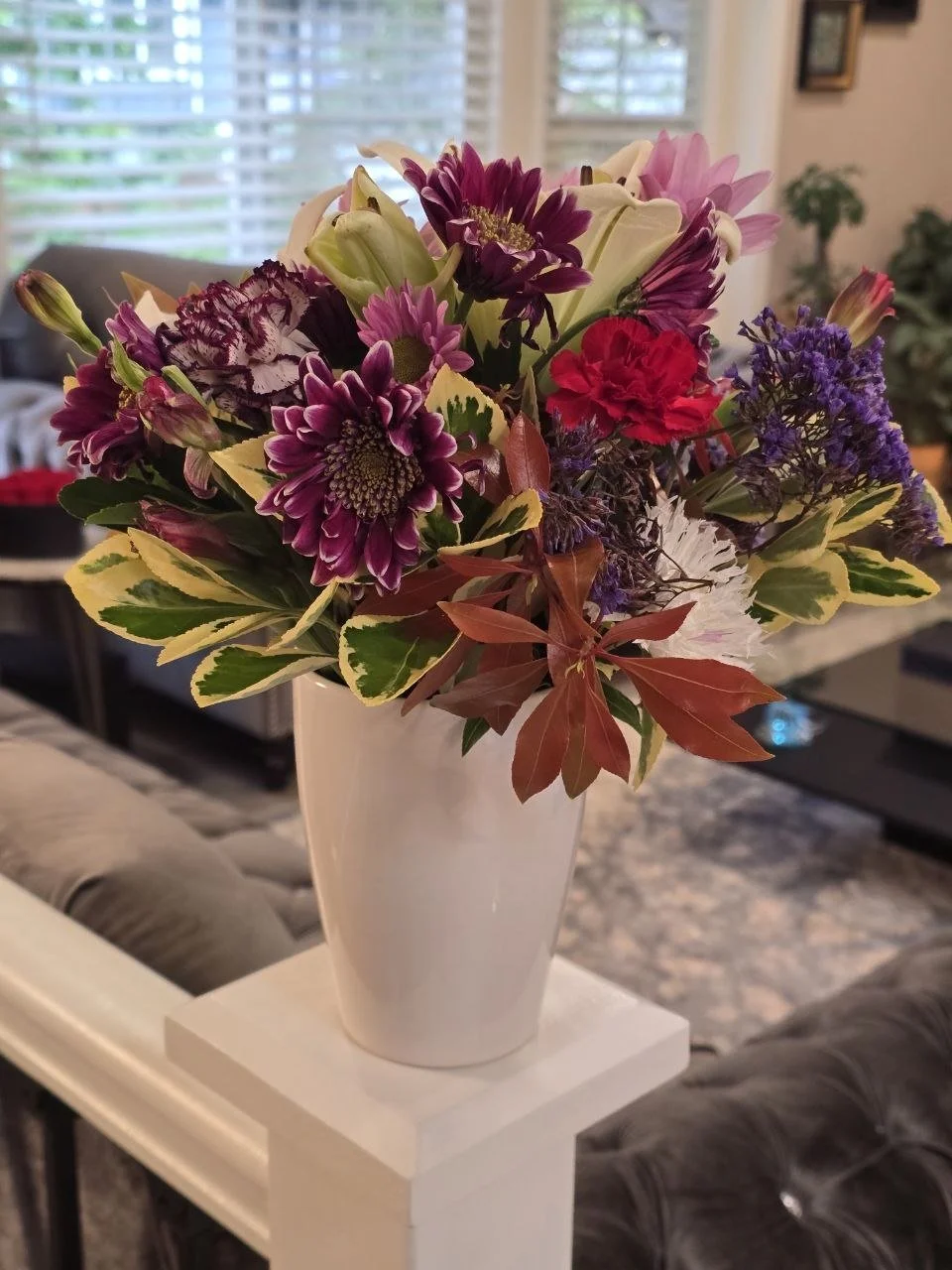 Colorful bouquet of mixed flowers in a white vase on a white table in a living room with large window and plants in the background.
