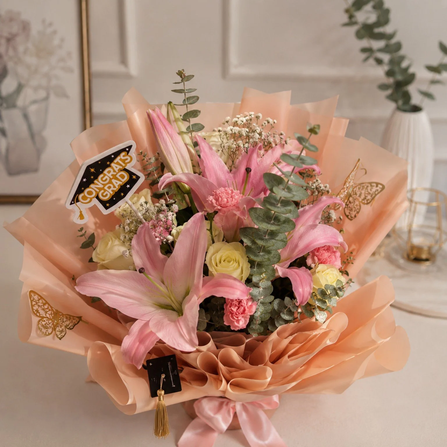 A bouquet of pink and white flowers wrapped in peach-colored paper with decorative butterflies, an 'Congrats Grad' tag, and a small gold tassel.