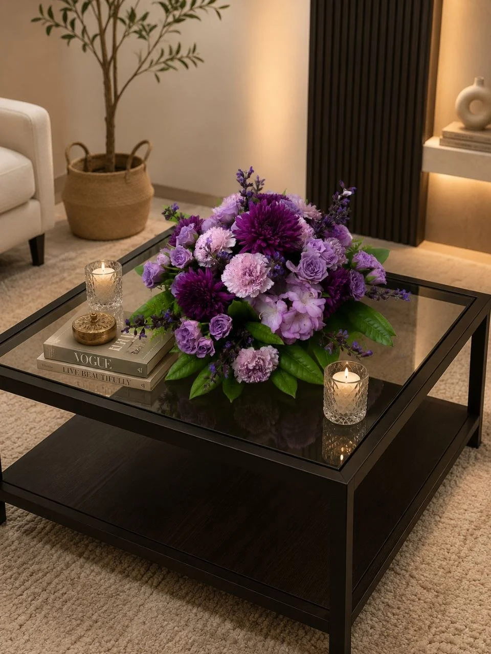 A living room with a black coffee table holding a large bouquet of purple and pink flowers, two lit candles in glass holders, and a stack of magazines. In the background, there is a white sofa, a potted plant, and wall shelves with decorative items.