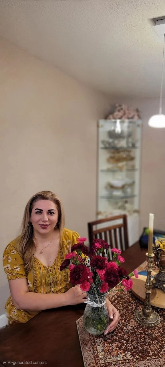 A woman with long blonde hair wearing a yellow patterned blouse, sitting at a dining table, holding a glass vase with pink and red flowers.