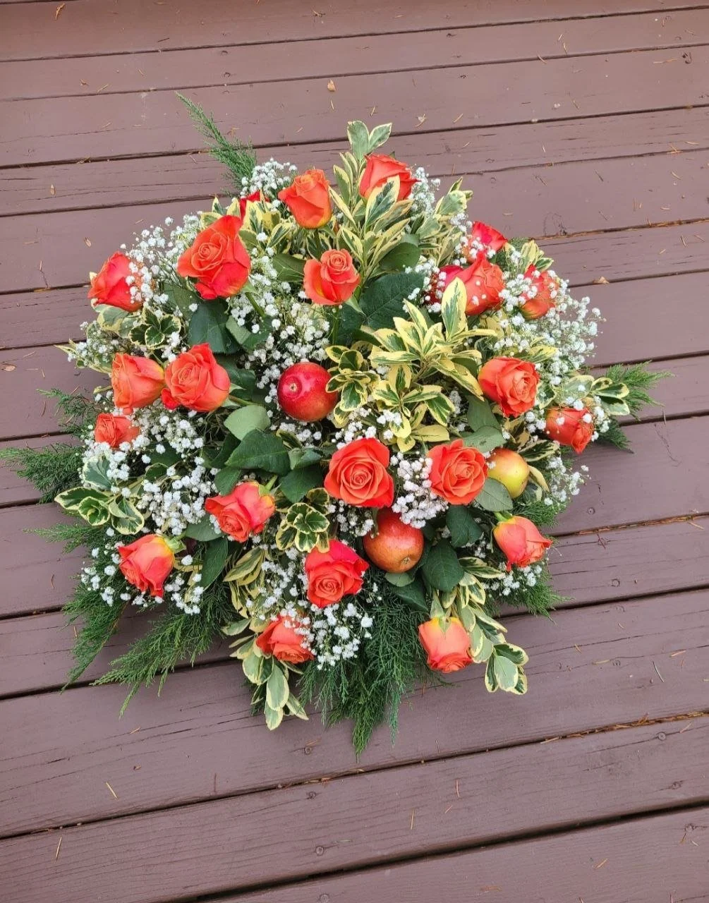 A floral arrangement with orange roses, green foliage, yellow and green variegated leaves, white baby's breath, and small red apples, placed on a wooden surface.
