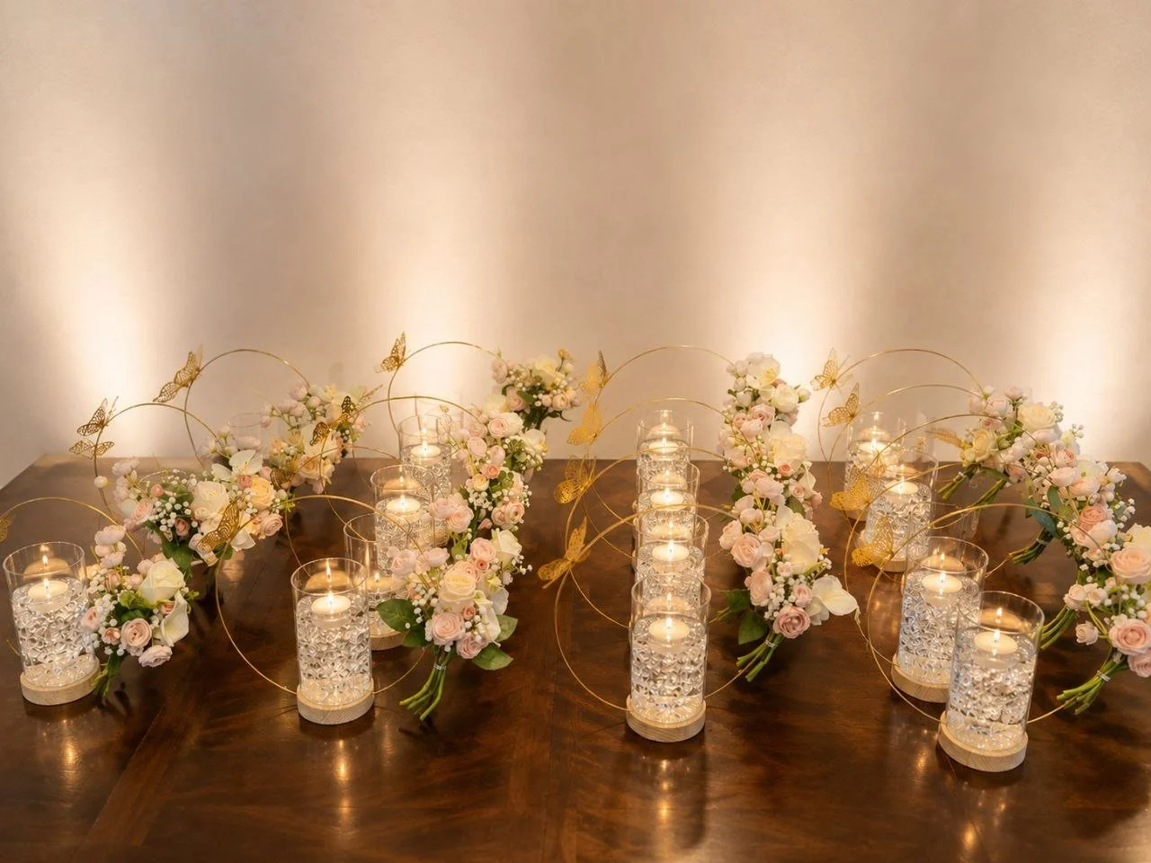 Decorative table centerpiece with candles, pink and white roses, greenery, and gold butterfly ornaments arranged in a symmetrical pattern.
