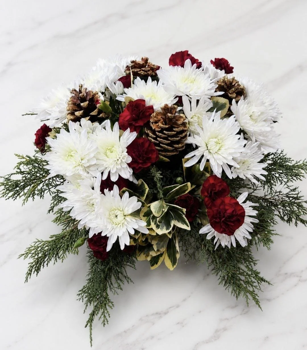 A Christmas floral arrangement with white chrysanthemums, red carnations, pine cones, and greenery on a white marble surface.
