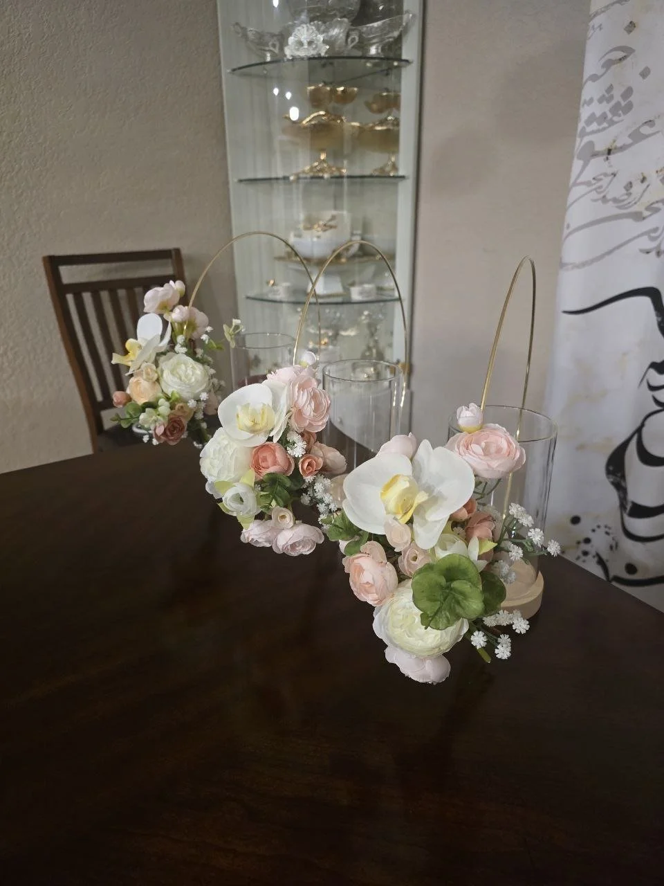 A floral centerpiece with white and pale pink flowers, including orchids, on a dark wooden table, with a glass display case filled with gold and white decorative objects in the background.