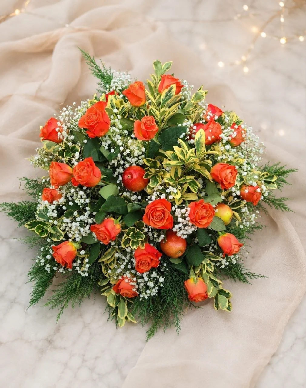 A floral arrangement with orange roses, small white baby's breath flowers, green ferns, and variegated leaves, placed on a light-colored, possibly marble, surface with soft beige fabric background.