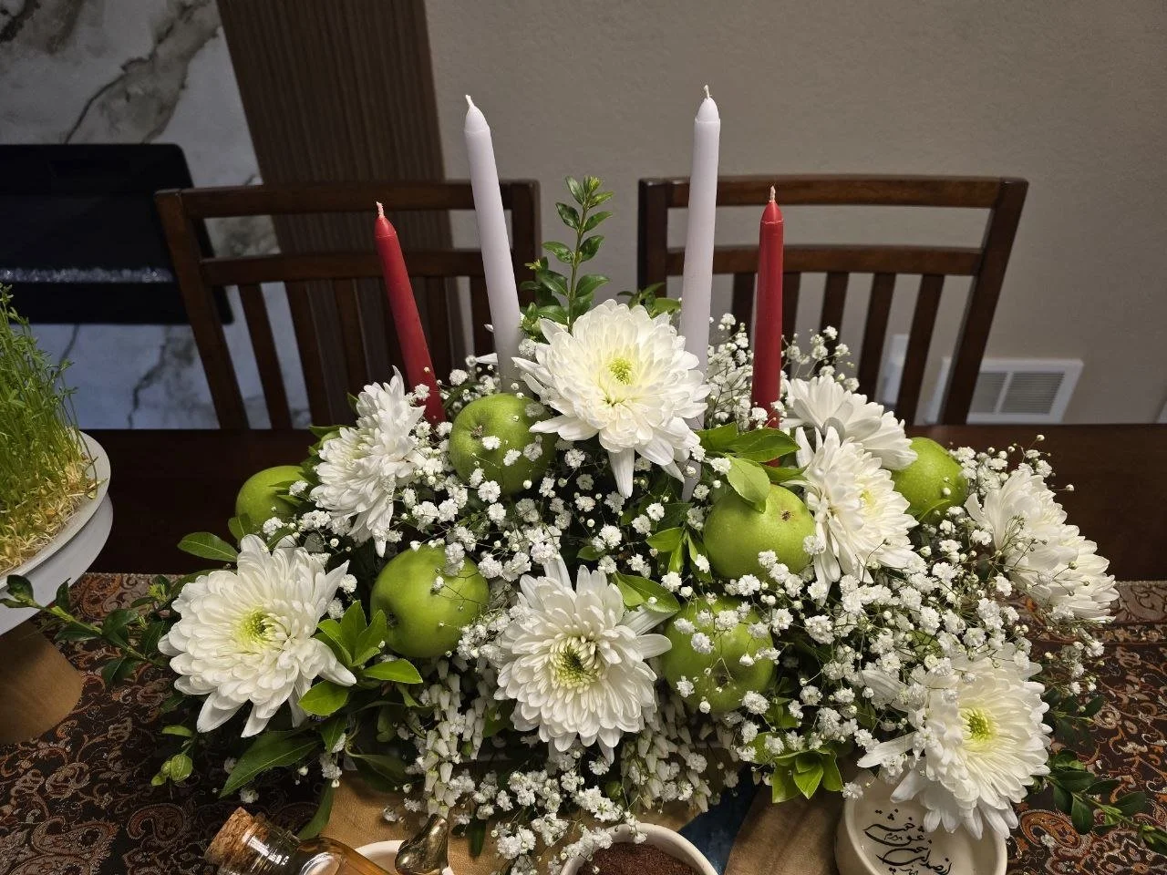 Colorful floral centerpiece with white flowers, green apples, and four candles on a dining table.