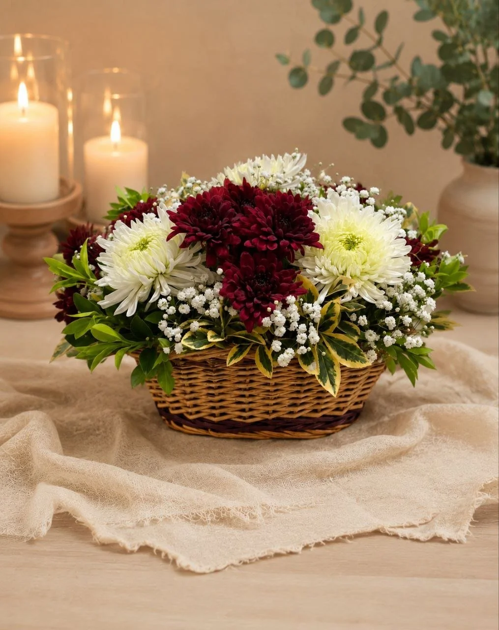 A woven basket filled with white, burgundy, and cream flowers, surrounded by green leaves, placed on a beige fabric tablecloth with candles and potted plants in the background.