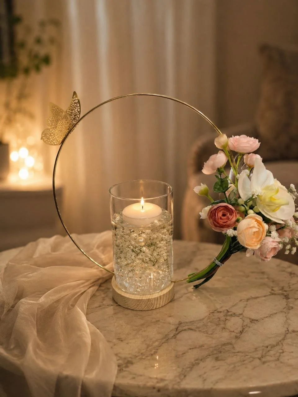 A lit candle in a glass holder with decorative pearls floats inside, placed on a marble table. A contemporary floral arrangement with pink, peach, and white flowers is positioned nearby. A sheer fabric drapes across the table.