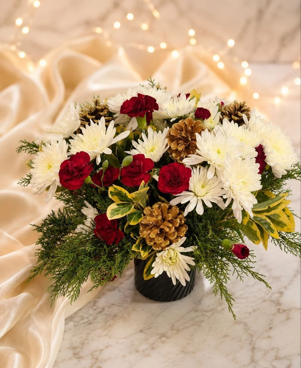 Holiday floral arrangement with white, red, and gold pinecones on a marble surface, with a soft gold fabric and fairy lights in the background.
