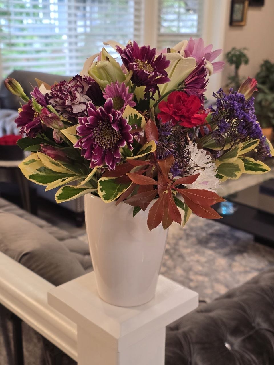 Vase filled with a colorful variety of fresh flowers including purple, pink, red, and white blooms, placed on a white stand in a living room with large windows in the background.
