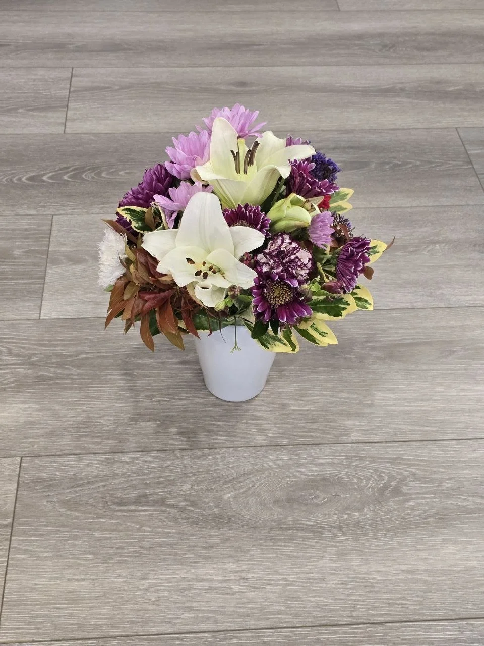 A bouquet of white, purple, and pink flowers in a white vase on a wooden floor.