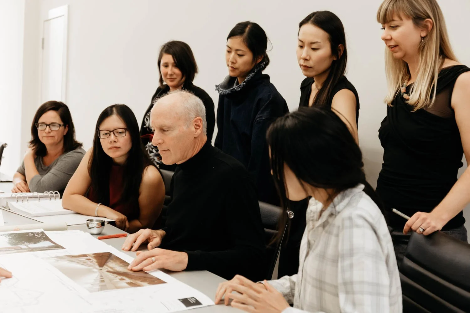 A diverse group of women and an elderly man are gathered around a table looking at large printed images and documents during a meeting, with some standing and others sitting.
