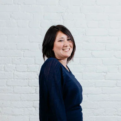 A woman with shoulder-length dark hair smiling and wearing a navy blue top, standing in front of a white brick wall.