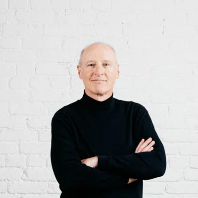 A middle-aged man with short white hair, wearing a black turtleneck sweater, standing with arms crossed in front of a white brick wall.