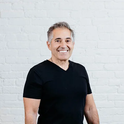 Smiling man with gray hair wearing a black T-shirt standing against a white brick wall.