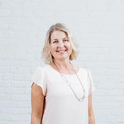 A smiling middle-aged woman with short, wavy blonde hair standing in front of a white brick wall, wearing a white blouse and a long necklace.