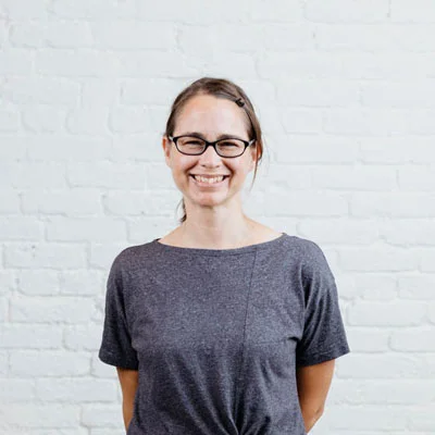 A woman smiling, wearing glasses and a dark gray t-shirt, standing in front of a white brick wall.