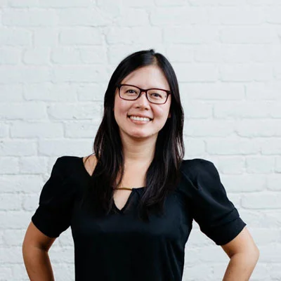 A woman with glasses and long dark hair, smiling and wearing a black top, standing in front of a white brick wall.