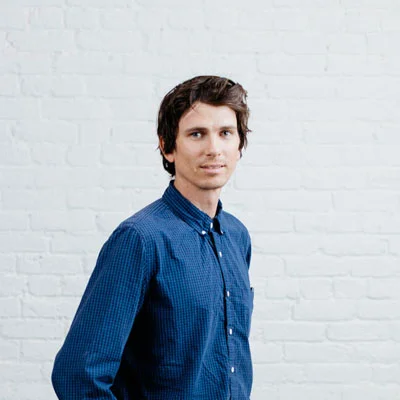 Man with dark hair wearing a blue checkered shirt standing against a white brick wall