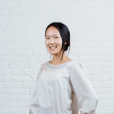 Woman smiling in front of a white brick wall, wearing a light-colored blouse with decorative details around the collar.