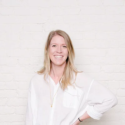 A woman with blonde hair smiling, dressed in a white shirt, standing in front of a white brick wall.