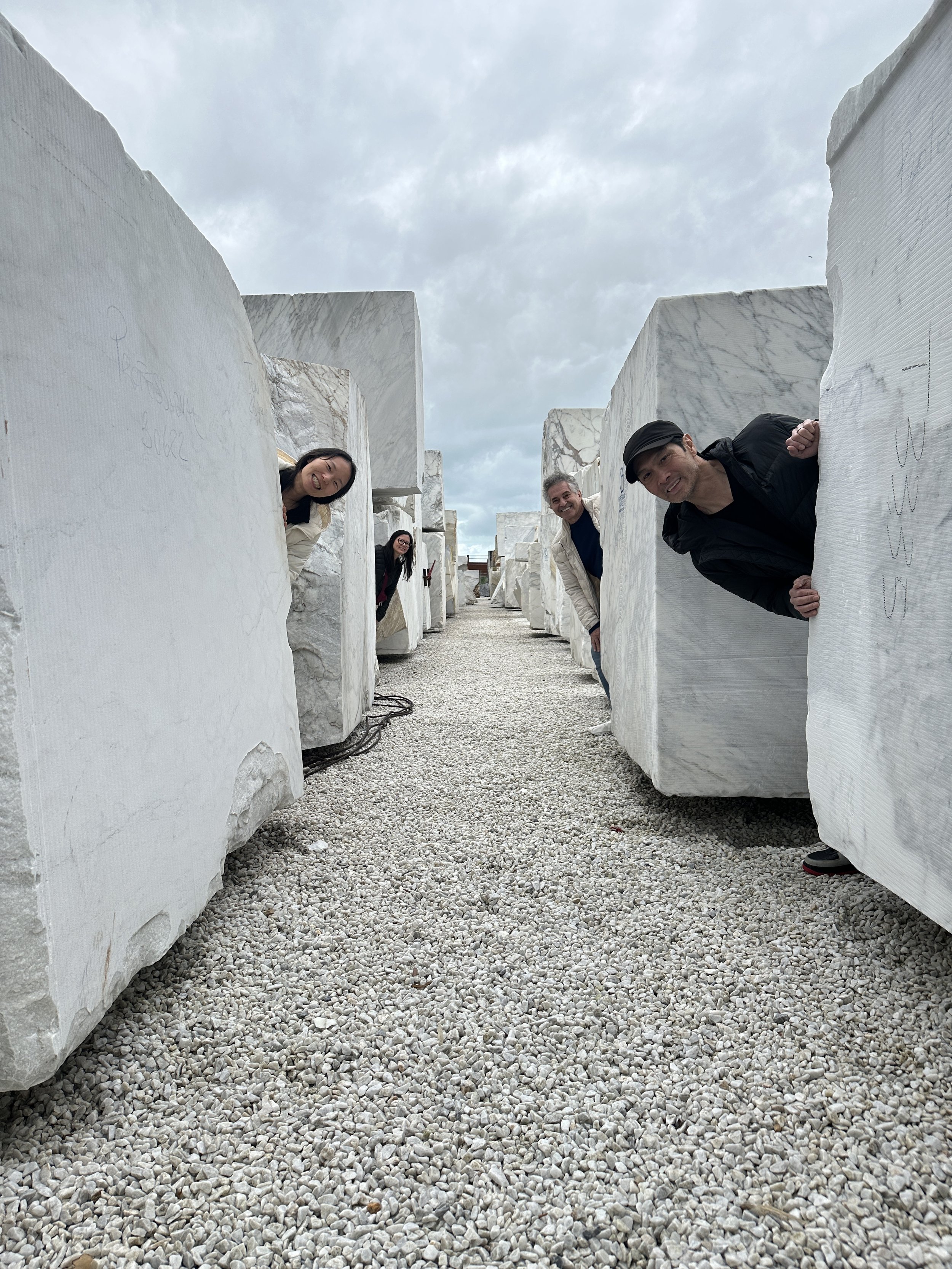 Four people peeking out from behind large white marble blocks in an outdoor sculpture park with cloudy sky.