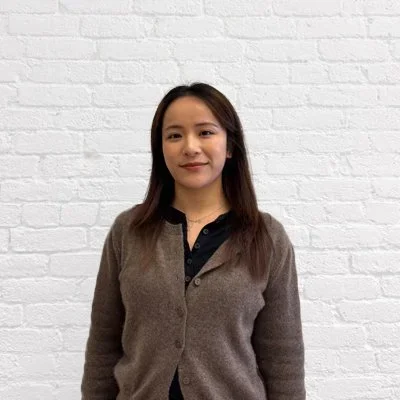 A woman with long dark hair wearing a brown cardigan over a black shirt stands against a white brick wall.