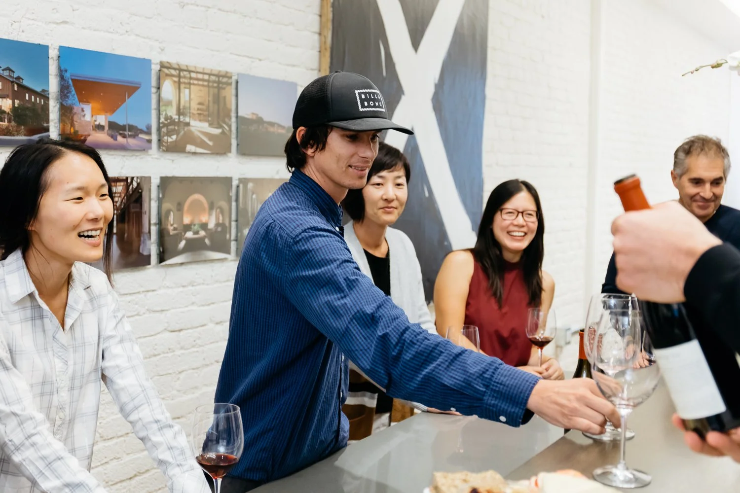 Group of people at a bar or social gathering, with some holding glasses of wine, smiling and engaging with a person pouring wine from a bottle.
