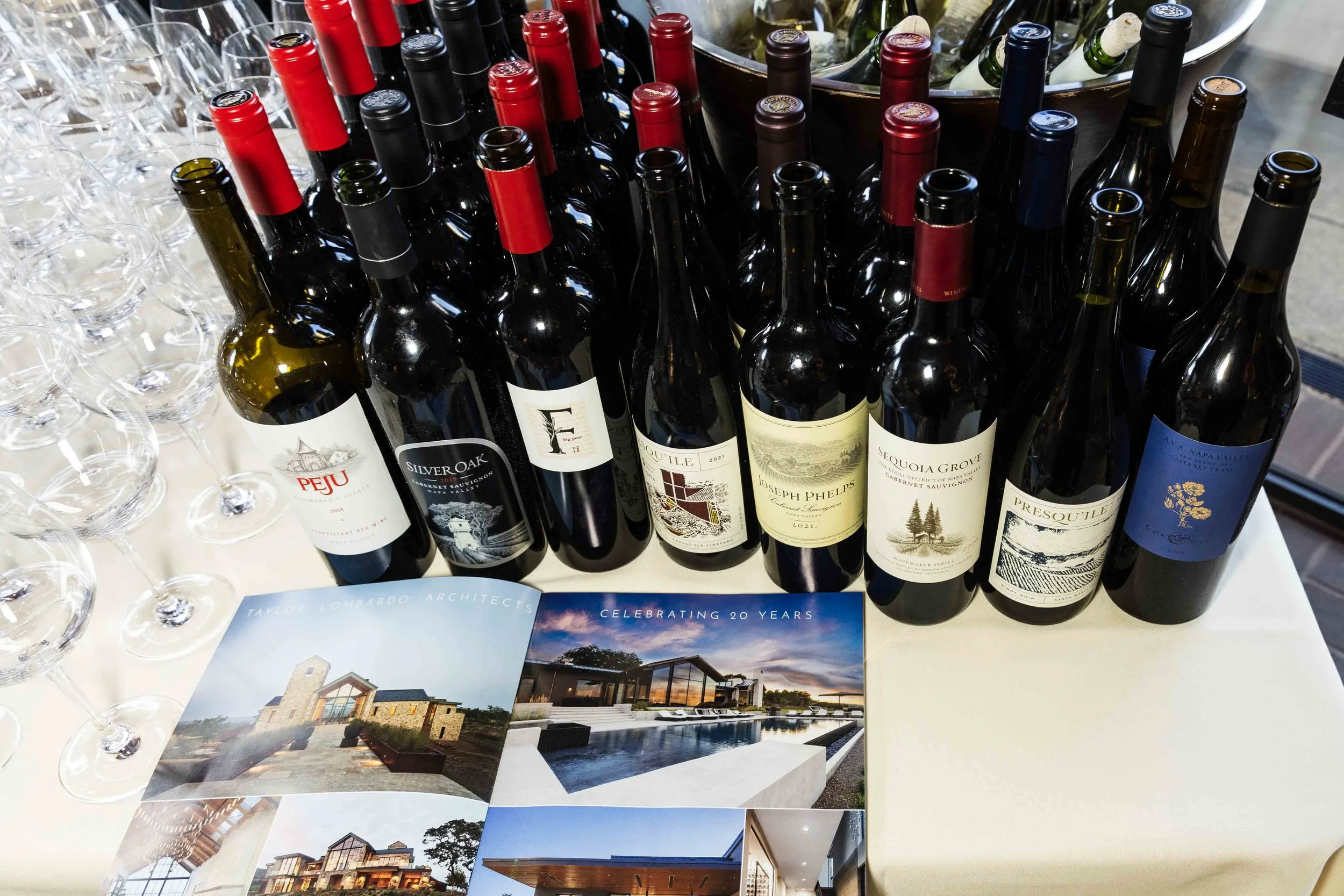 A display of various wine bottles with labels, some with red and black foil tops, next to empty wine glasses. Brochures or flyers showcasing architectural designs and modern homes are spread out on the table.