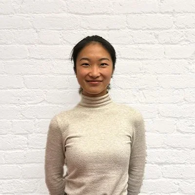 A woman with black hair in a ponytail smiling in front of a white brick wall, wearing a light-colored turtleneck sweater.